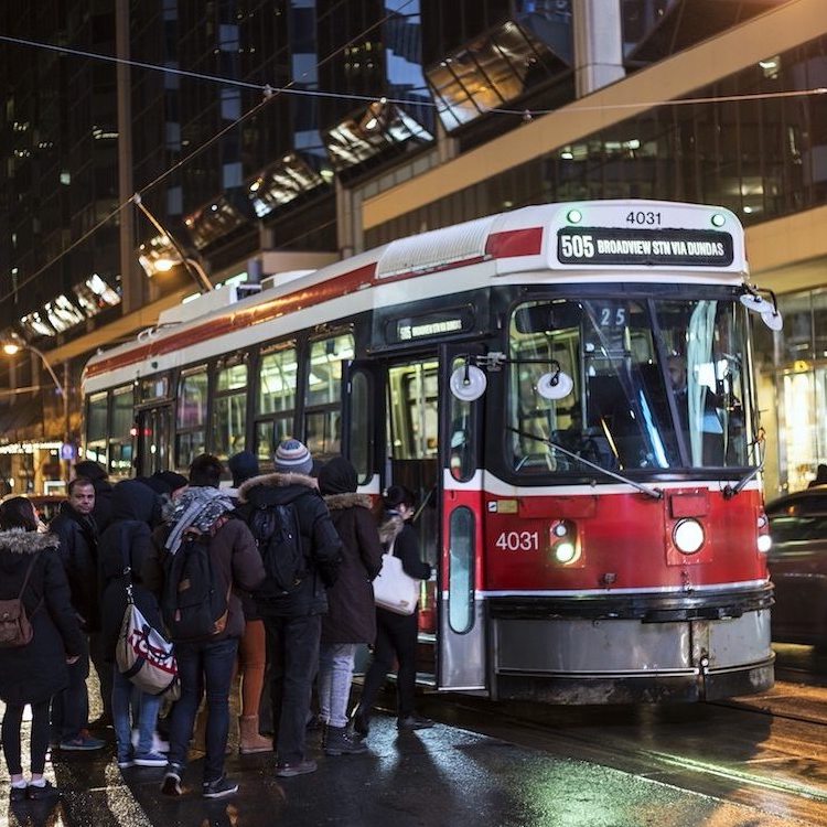 A large group of people waiting to board a streetcar on a busy nighttime steet.